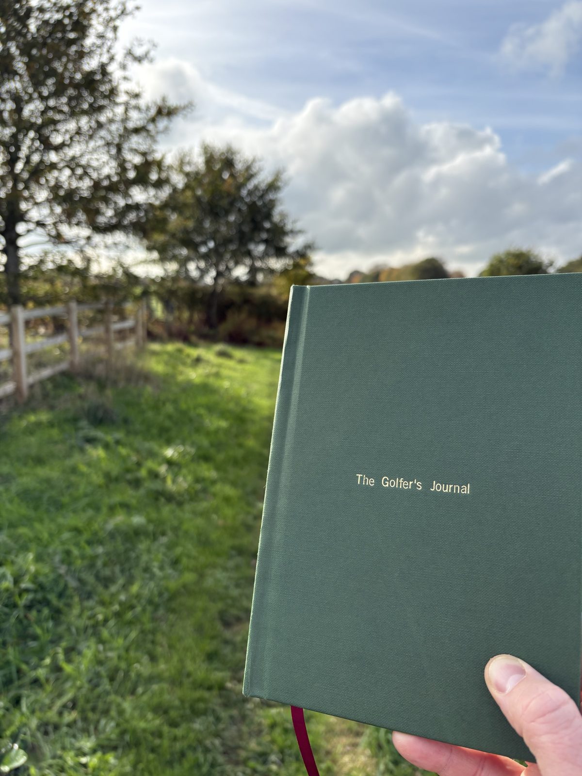 The Golfer's Journal held up beside a wooden fence on the course, under an autumn sky